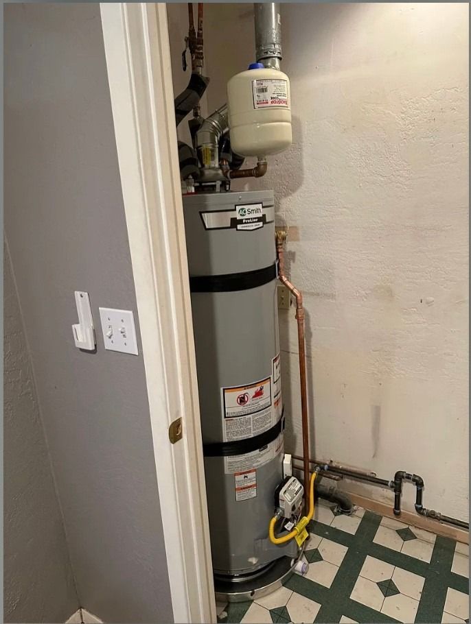 Water heater with expansion tank in a narrow utility closet, with exposed pipes and tiled floor.