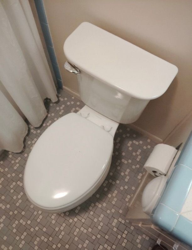 White toilet in a small bathroom with gray mosaic tile floor and a blue-tiled tub edge.