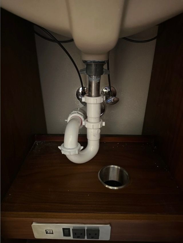 Under-sink plumbing with a white P-trap and drain pipe under a vanity, with a floor access opening nearby.