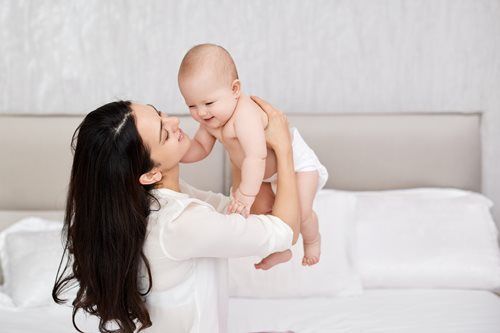 Woman holding and smiling at a baby in a diaper; on a bed in a light-colored room.