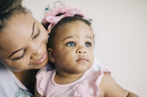 Woman embraces a baby with a pink headband and dress; both look away with gentle expressions.