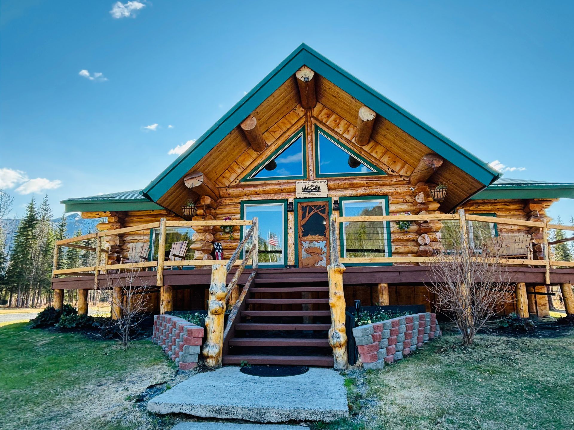 Log cabin with green trim and a deck, steps leading to the front door, blue sky in background.