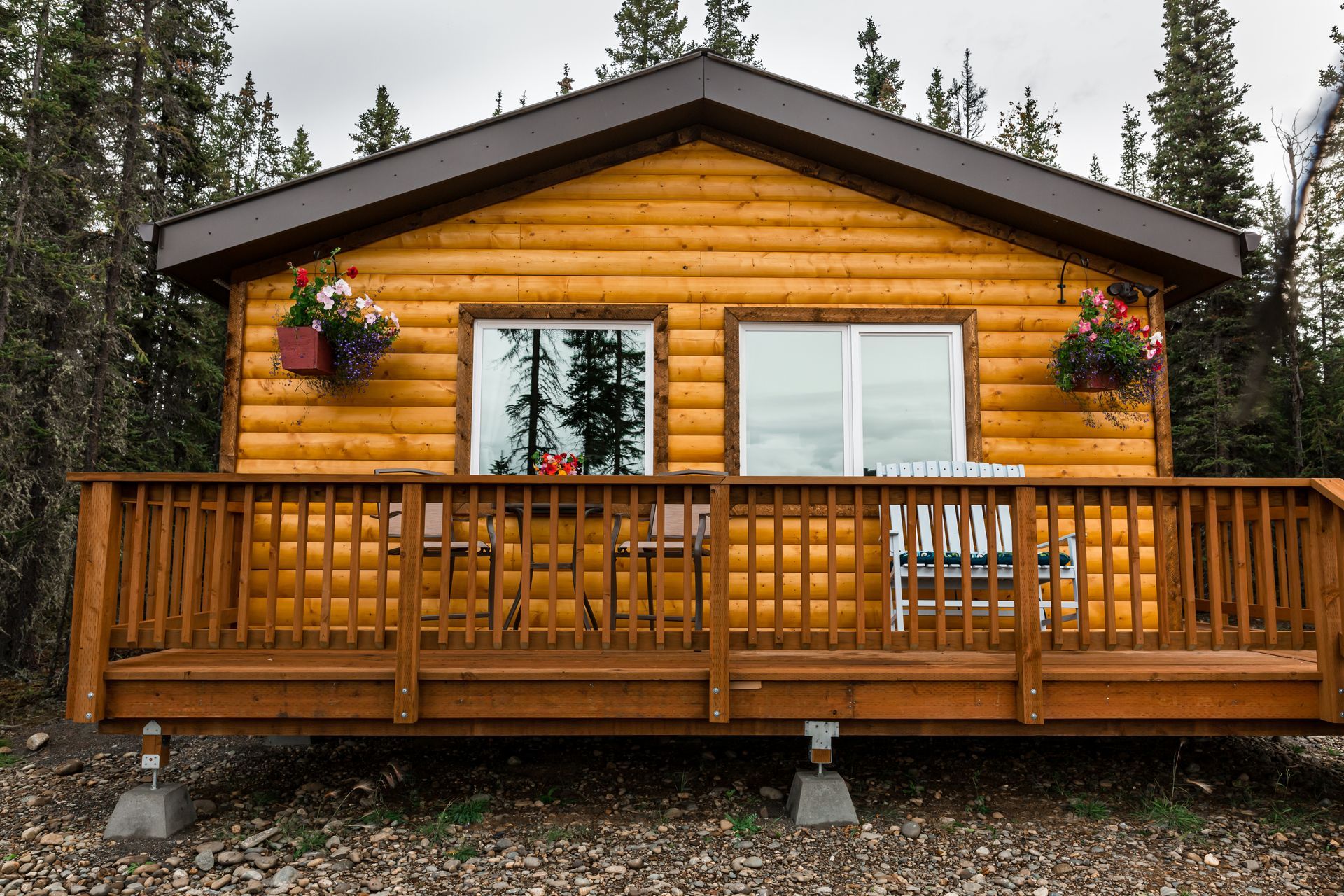 Wooden cabin with brown roof and deck, two windows, flower boxes, and a forest backdrop.