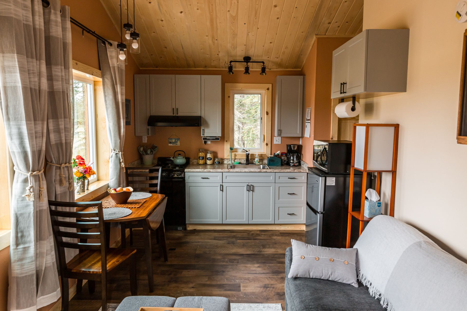Cozy, compact kitchen with gray cabinets, small appliances, and a table near a window with sheer curtains.