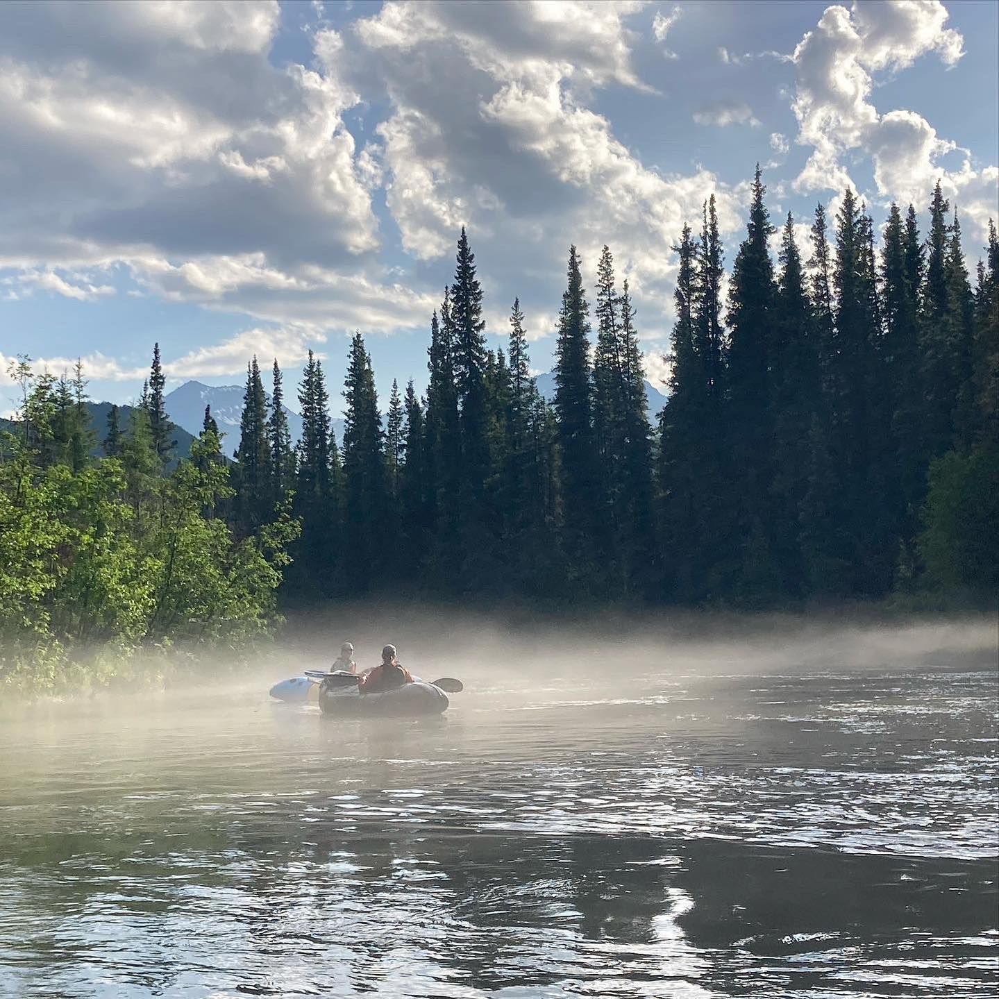 Two people in a small boat on a misty lake with tall pine trees and a cloudy sky.