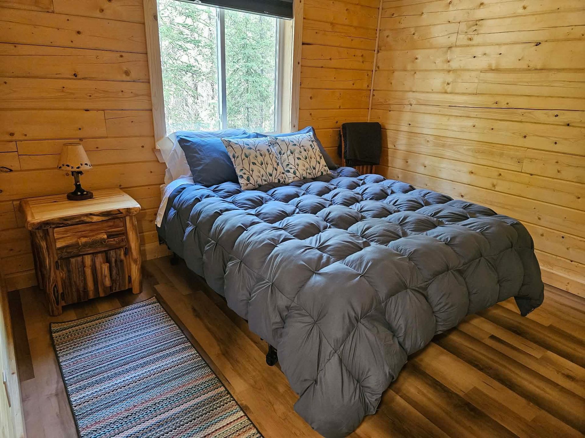 Cozy wood-paneled bedroom with a gray bedspread, striped rug, nightstand, and window view