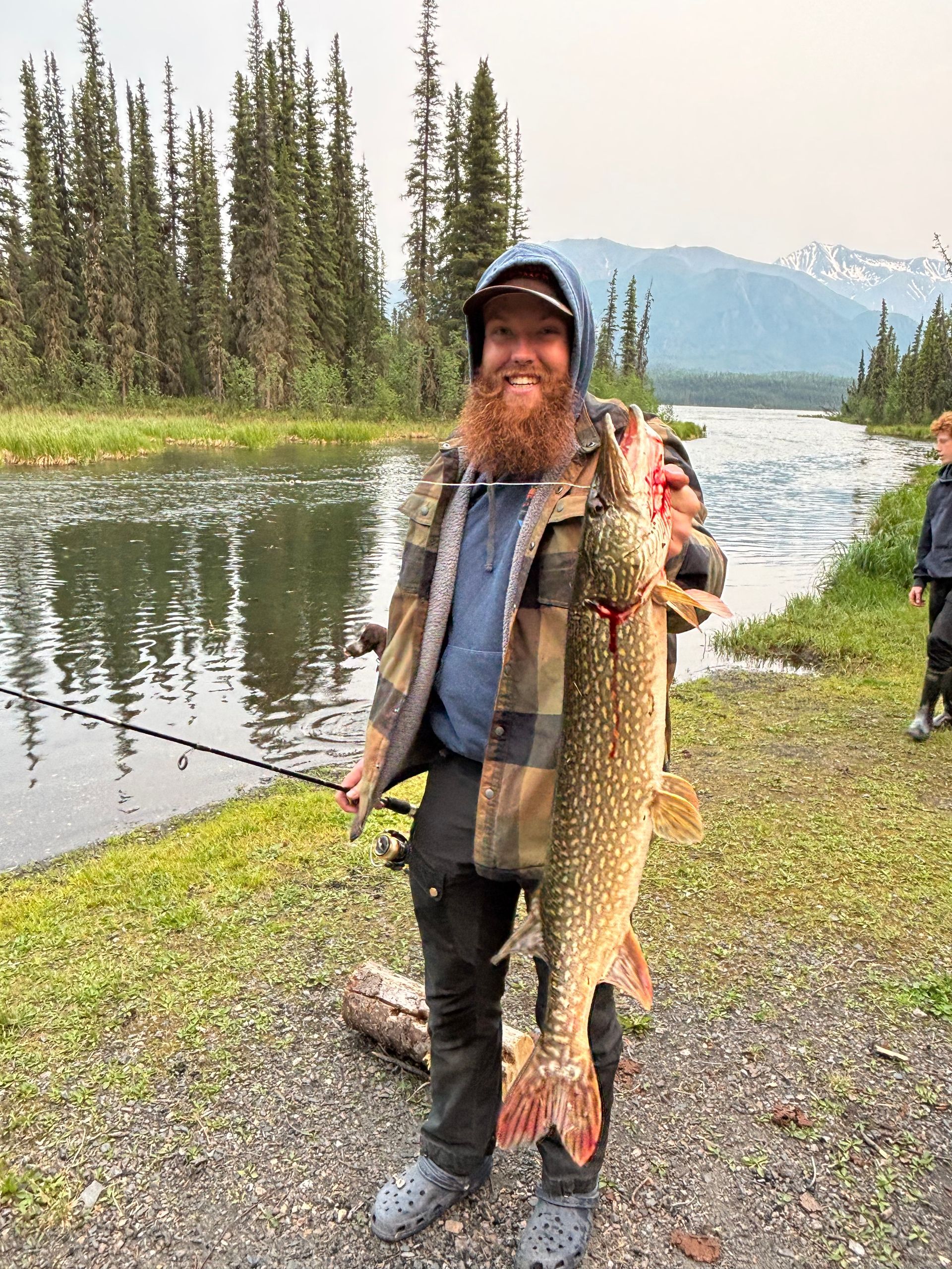 Man with red beard holding a large fish near a river with trees and mountains.