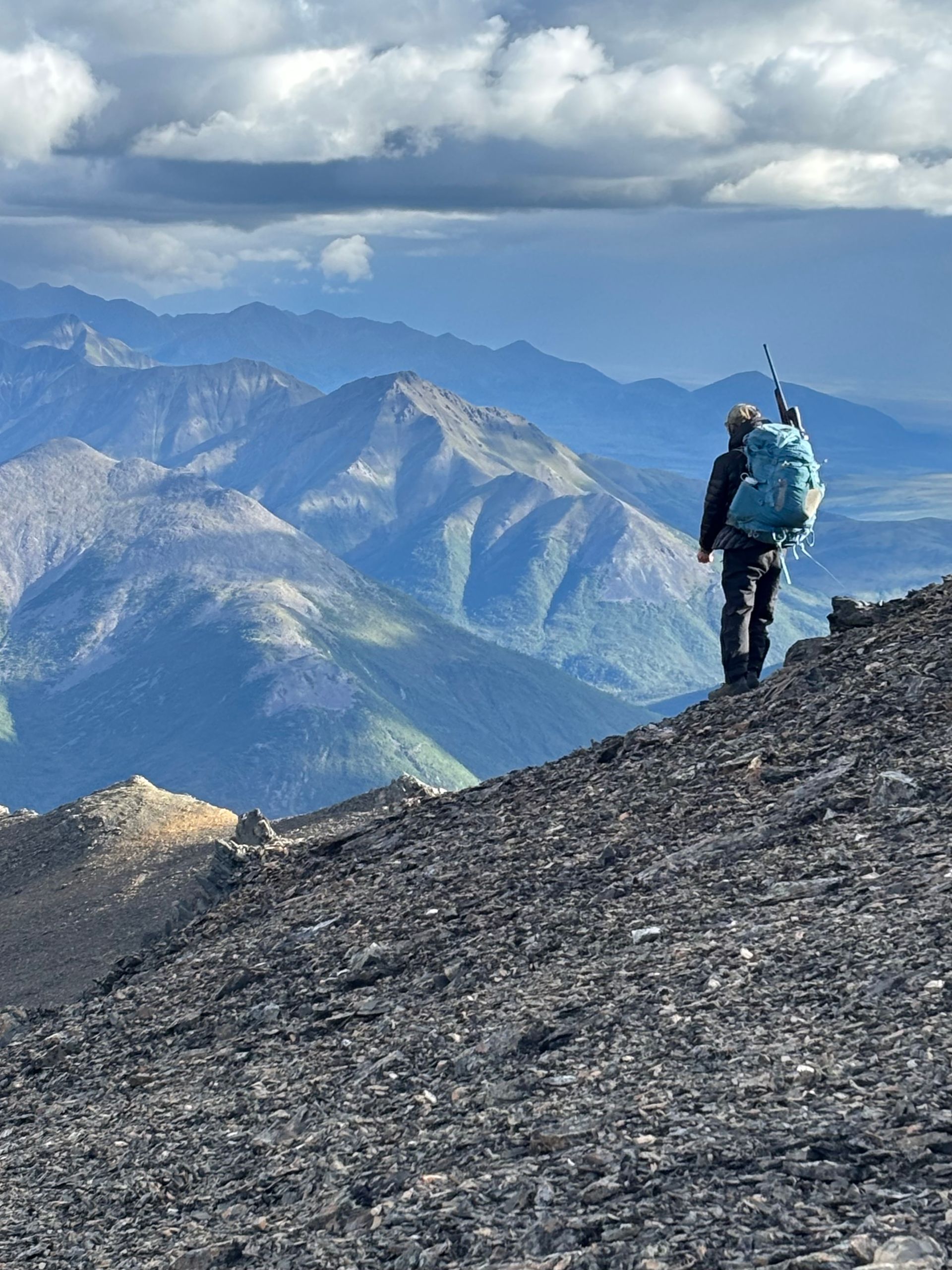 Hiker with backpack ascends a rocky slope, overlooking a mountain range under a cloudy sky.