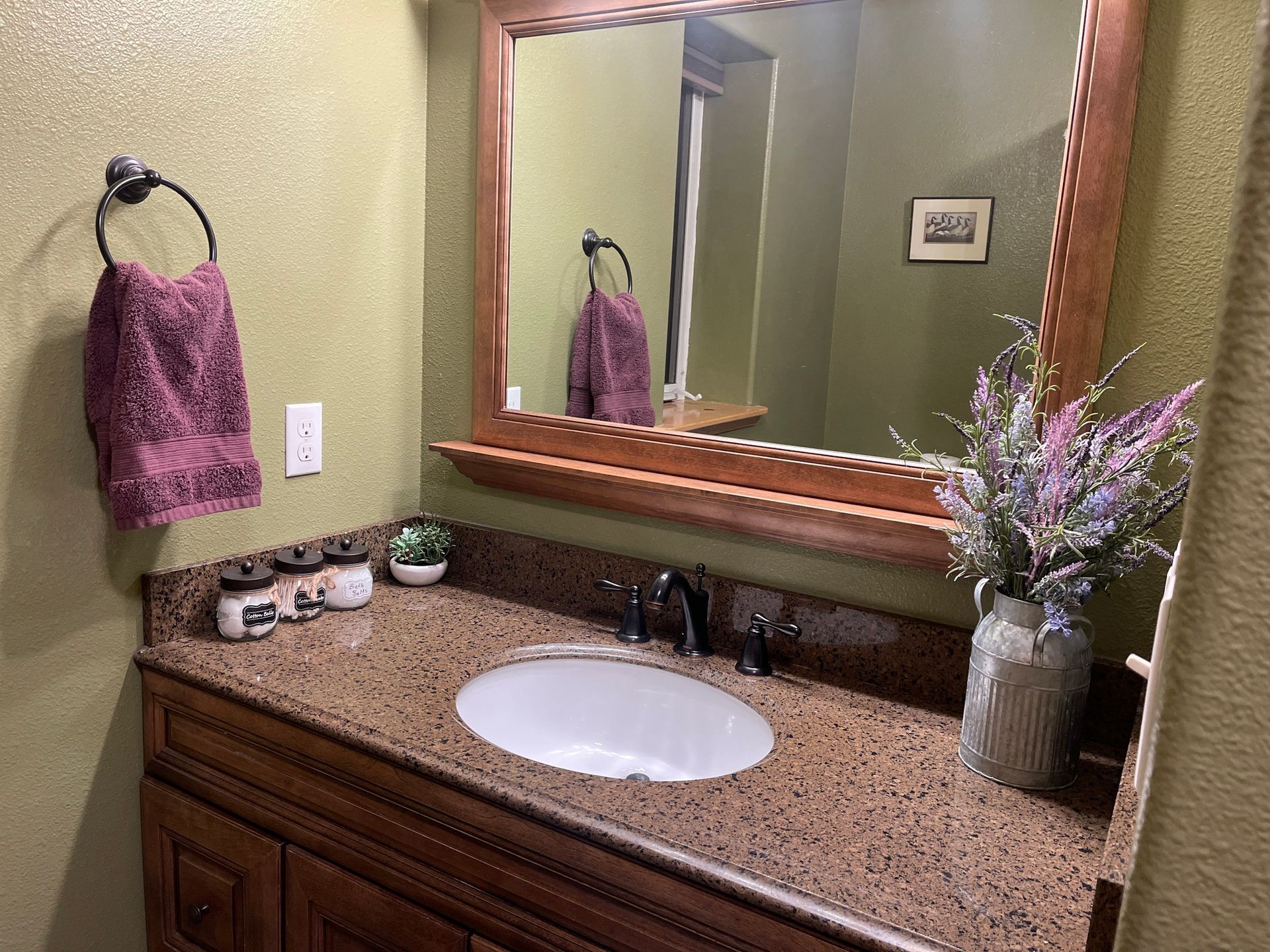 Bathroom vanity with oval sink, brown countertop, wooden cabinets and mirror, towel, and flower arrangement.