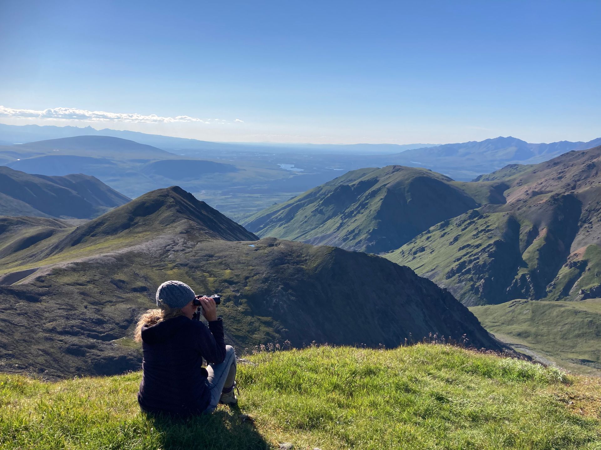 Woman seated on a grassy hilltop, using binoculars to view a mountain valley. Clear, sunny day.