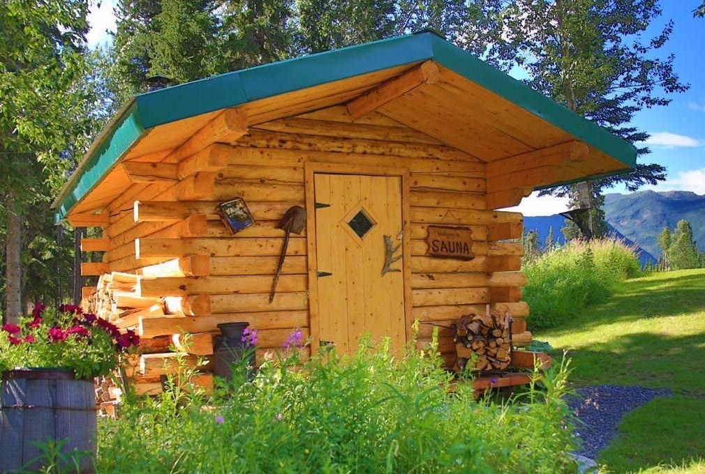 Small log cabin with a green roof and a wooden door, surrounded by greenery and flowers.