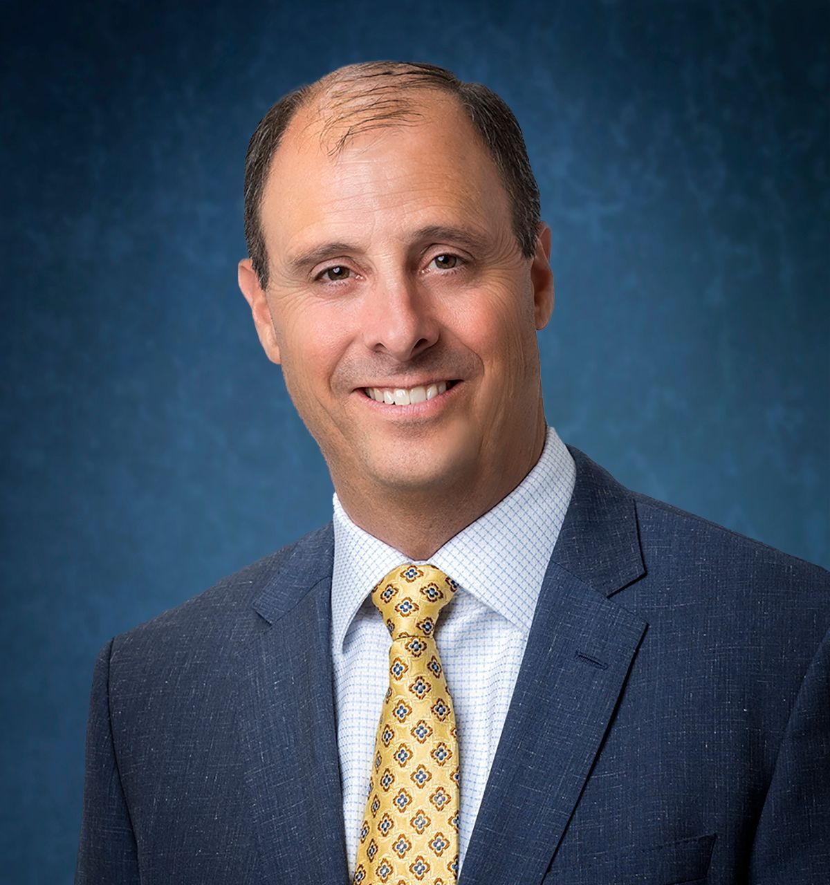 Man in blue suit, yellow patterned tie, smiling. Blue background.