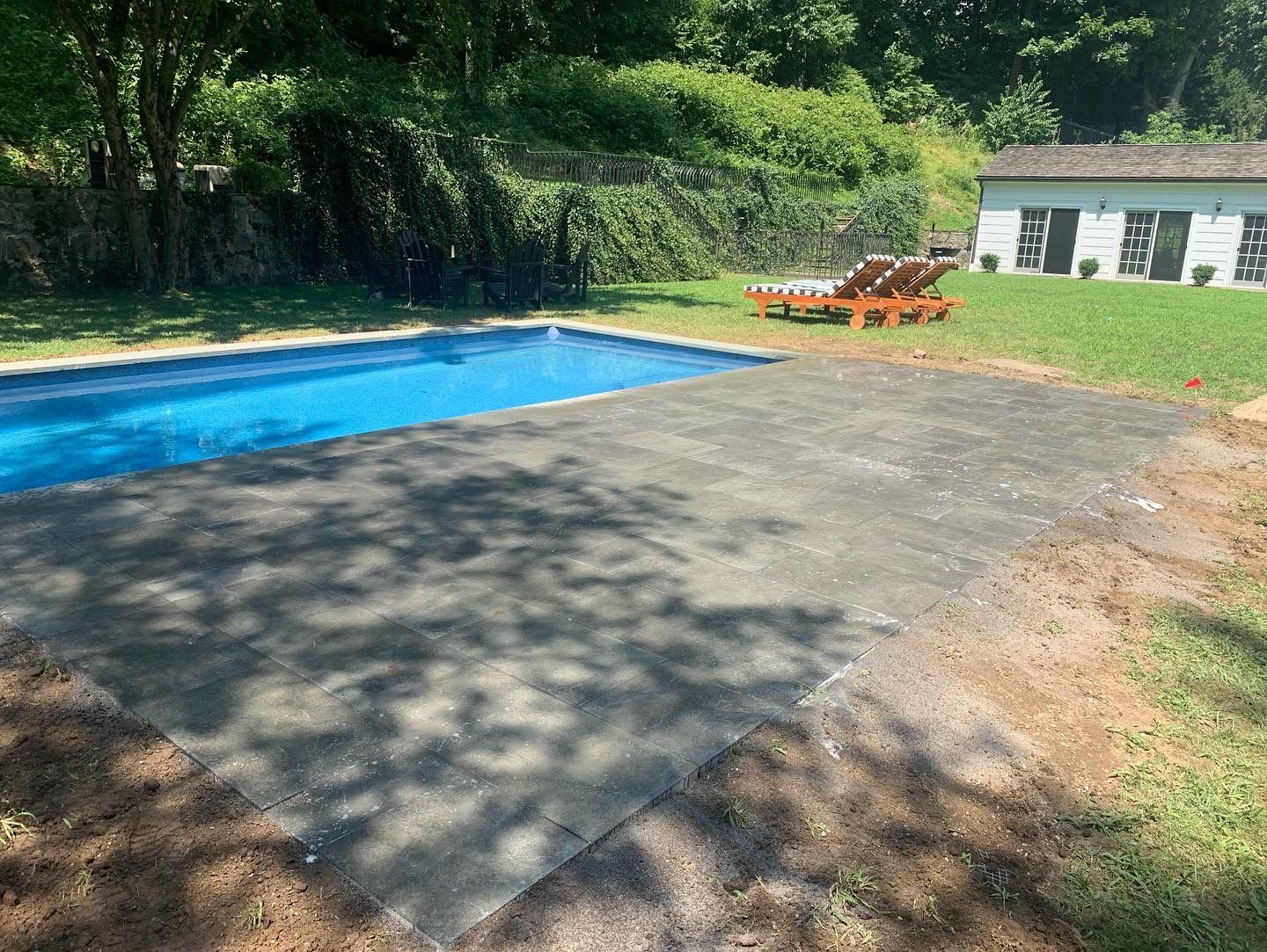 Backyard pool with blue water, paved deck, orange lounge chairs, and a white pool house by trees
