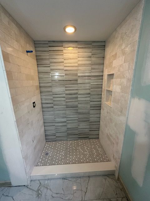 New tiled shower stall with gray accent wall, small shelf, and mosaic floor tile under a ceiling light