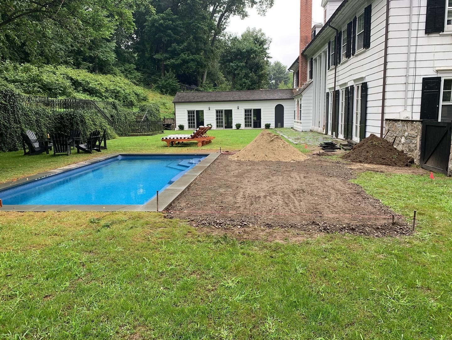 Backyard with a blue pool beside a white house and a large pile of dirt on the lawn.