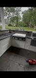 Outdoor patio with a built-in grill, stone counter, and green backyard trees beyond a concrete floor