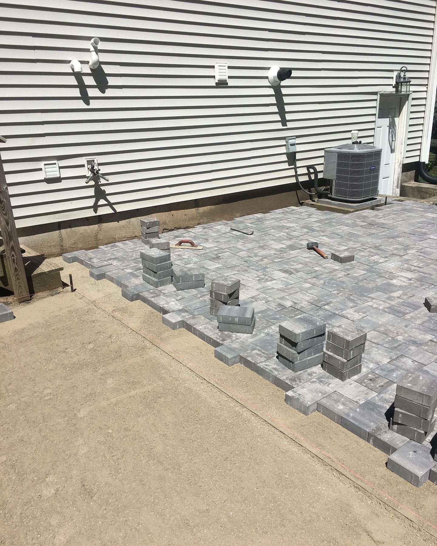Gravel patio under construction beside a house, with stacked pavers and tools against the siding.