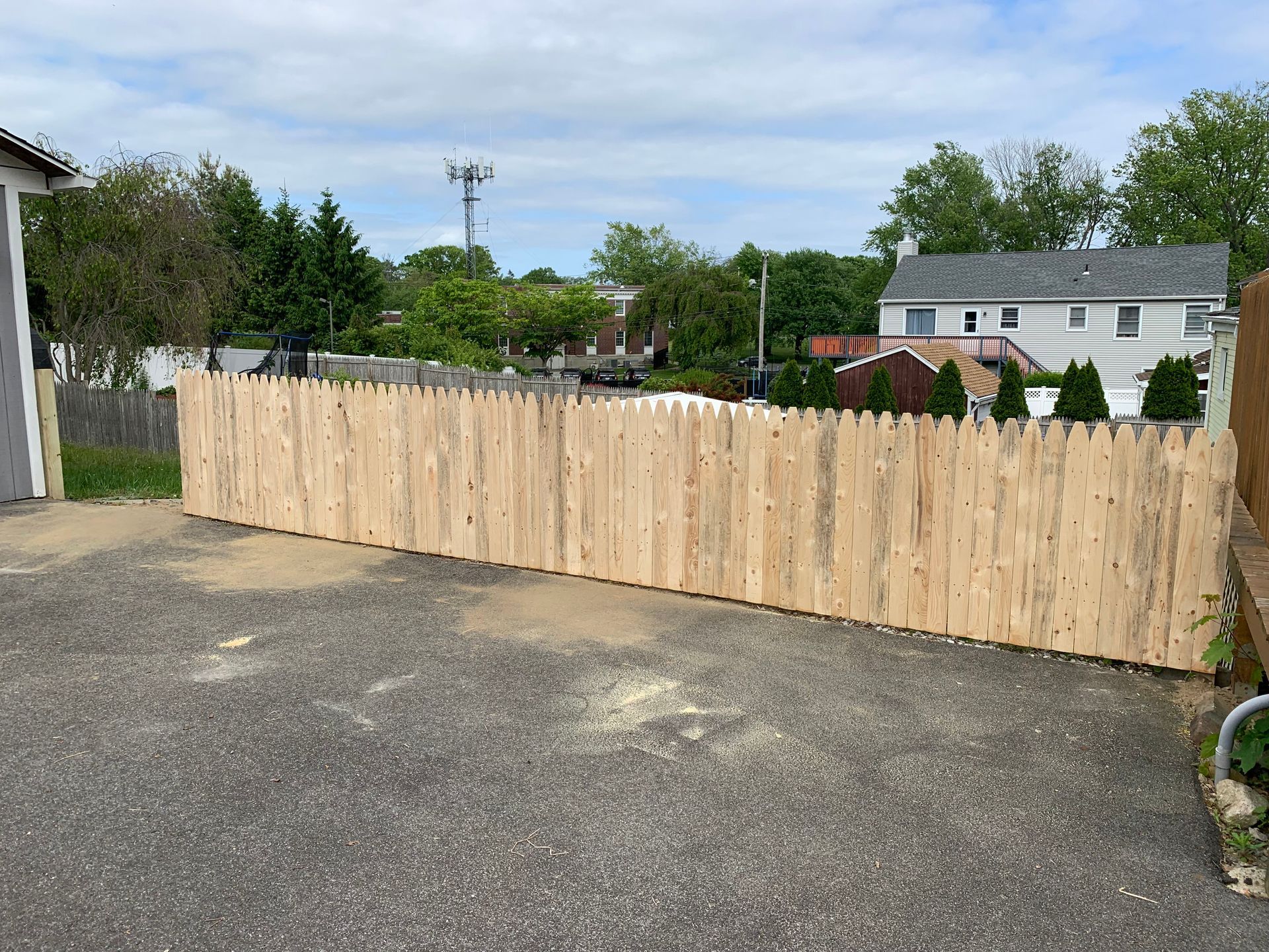Fresh wooden fence blocking a driveway beside a paved lot, with trees and houses in the background.