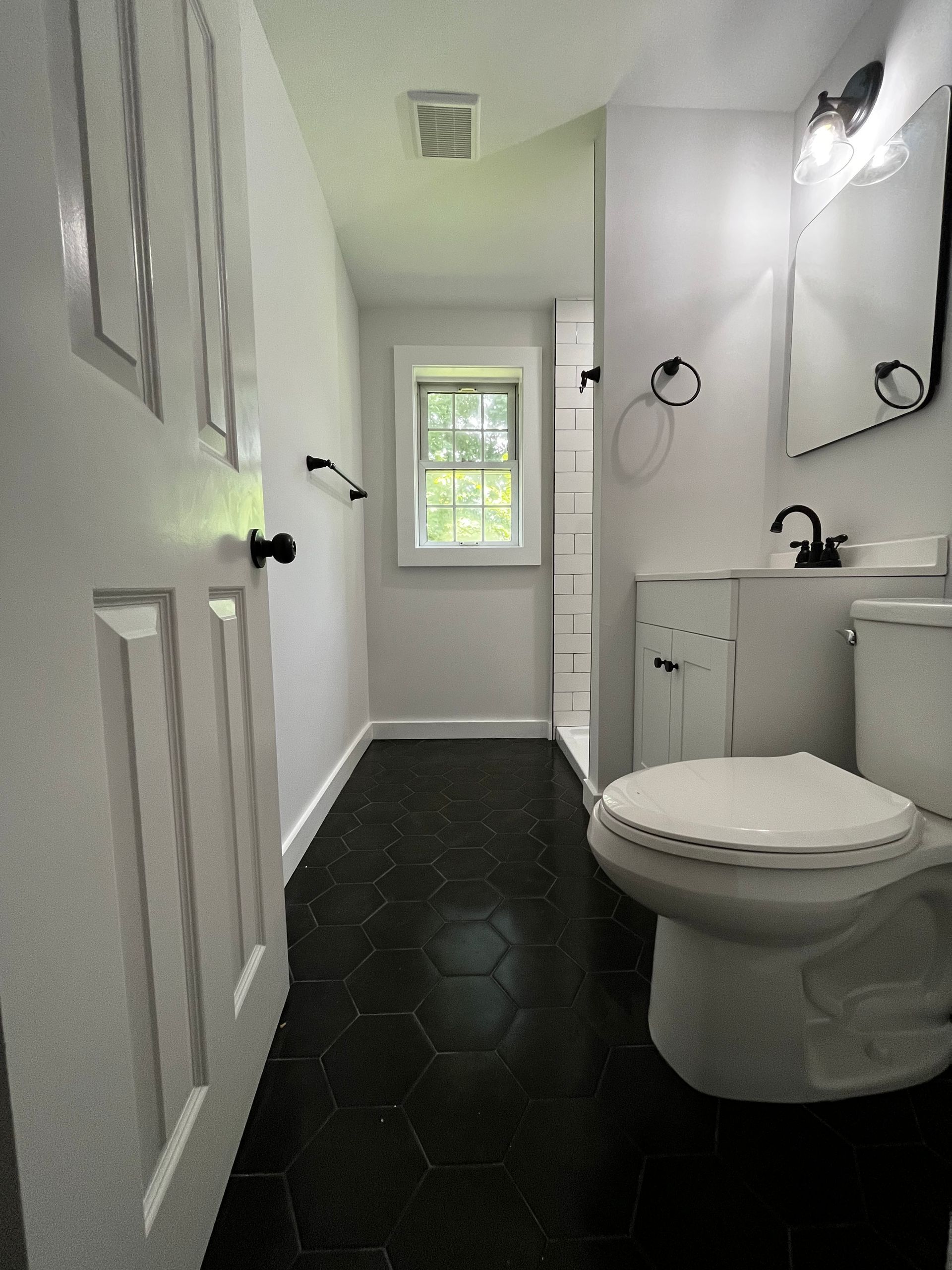 Narrow white bathroom with black tile floor, toilet, sink, mirror, and window at the far end