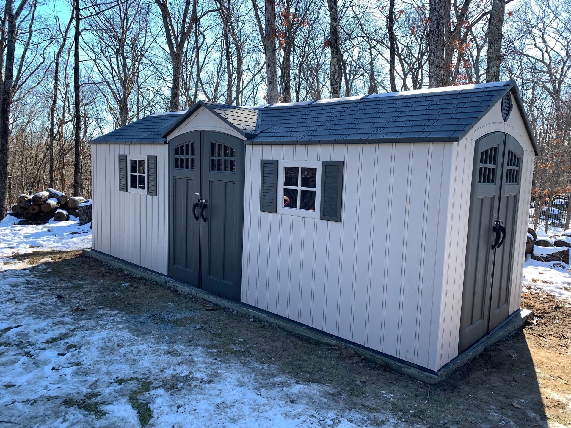 White backyard shed with double gray doors and windows, standing on snowy ground beside bare trees.