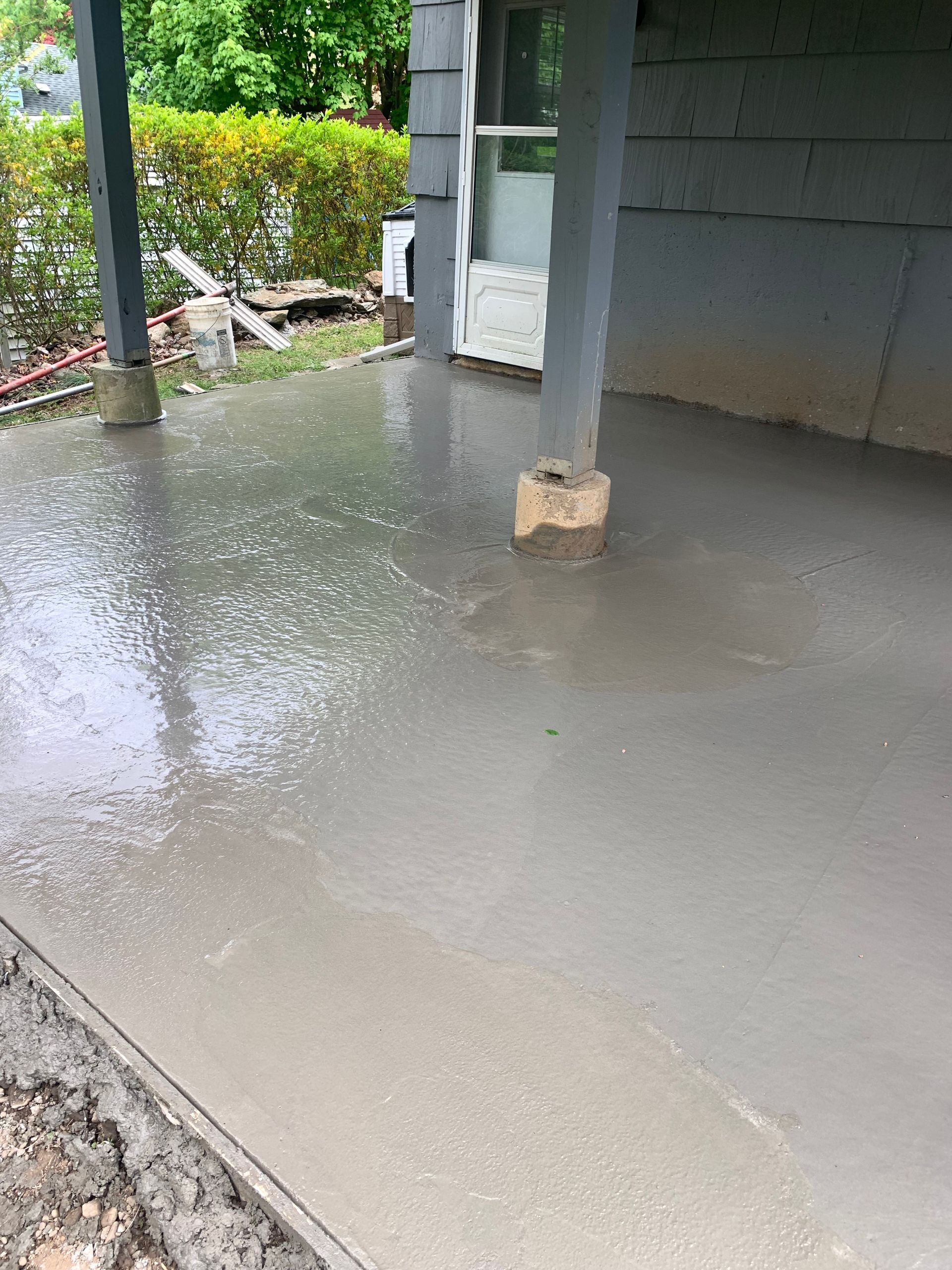 Freshly poured gray concrete porch with support posts and a wet garden area nearby