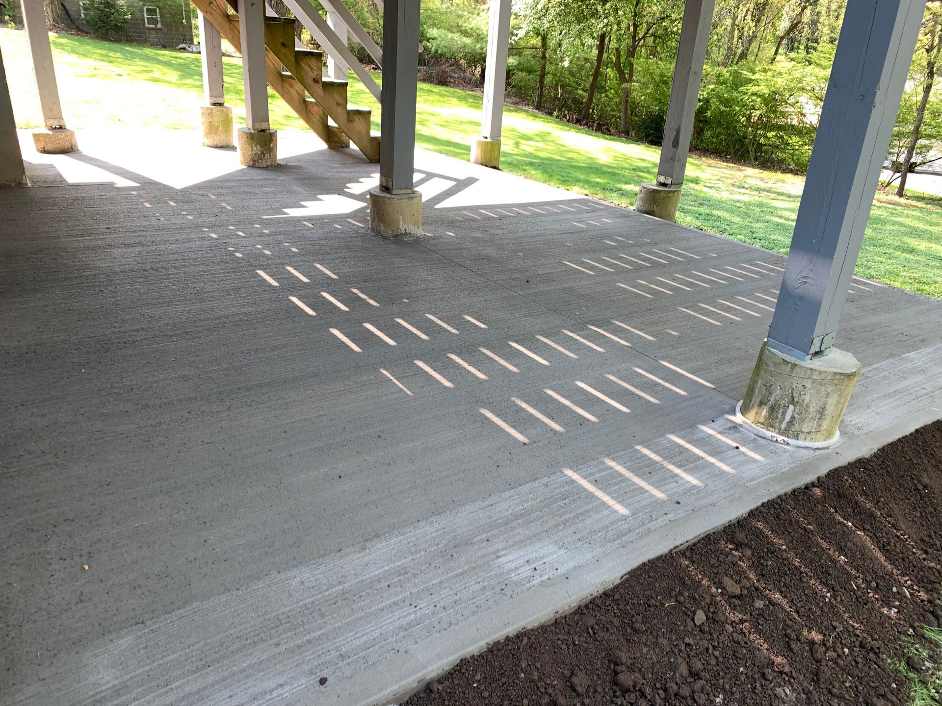 Concrete porch with metal posts casting striped shadows on the floor beside a gravel path and green lawn.