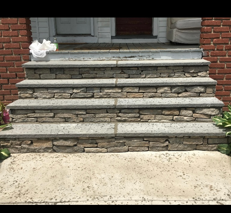 Stone front steps leading to a house entrance, with a brick wall and concrete walkway.