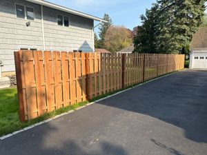 Wooden fence along a paved driveway beside a light gray house, with trees in the background.