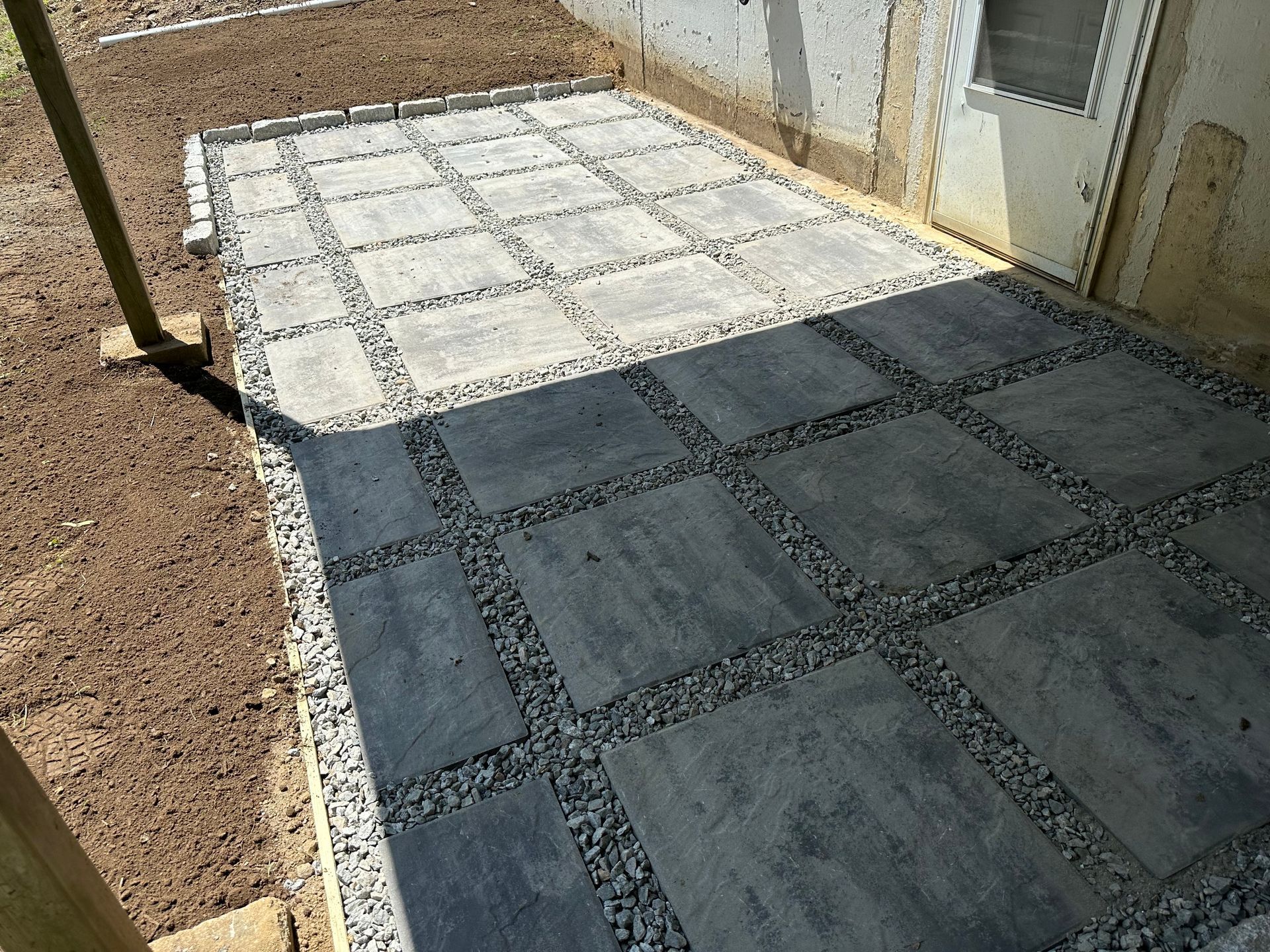 Concrete patio with square paver pattern beside a house, partly in sunlight and partly in shadow.