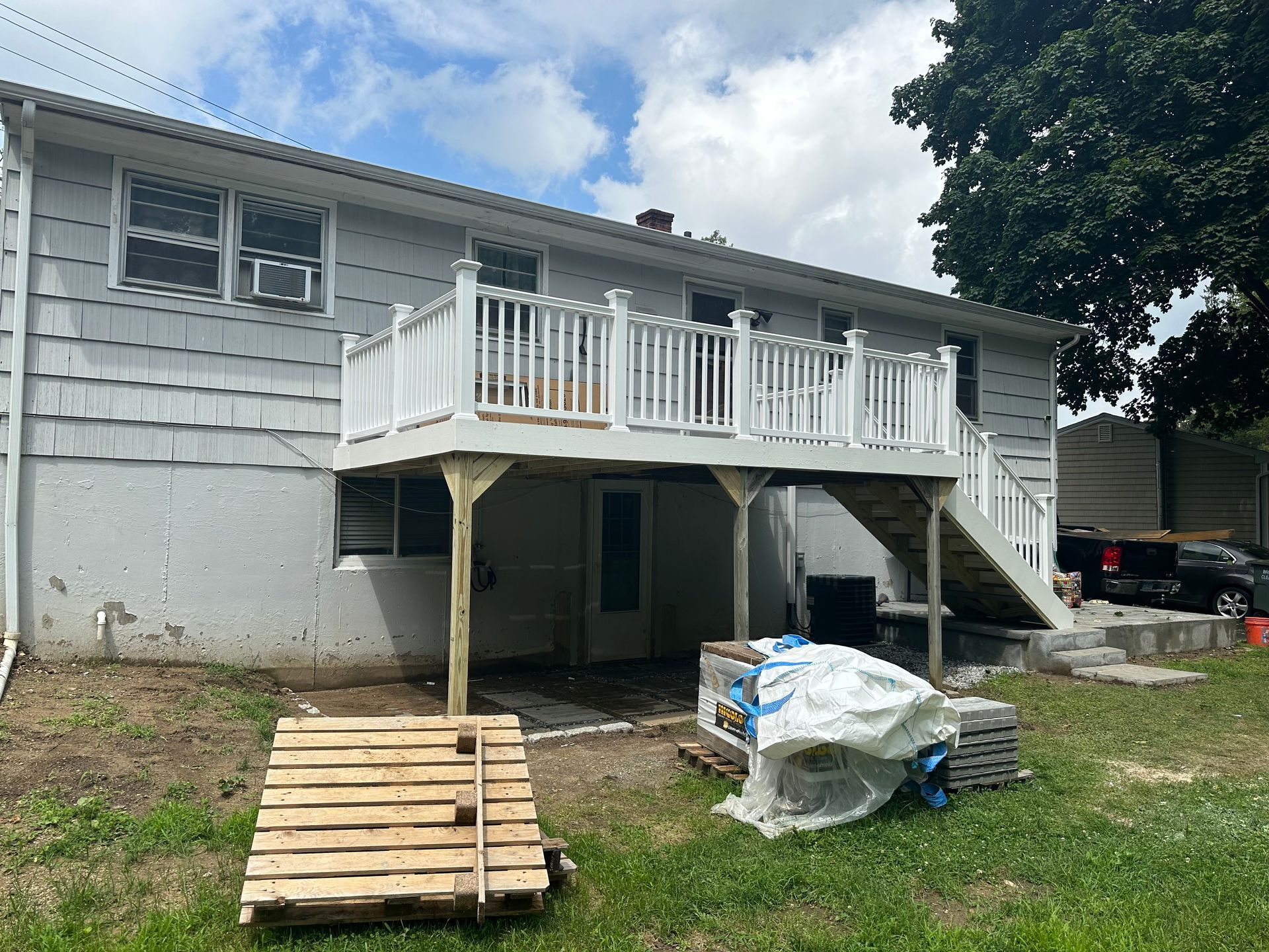 Exterior of a gray house with white balcony, stairs, and wooden pallet ramp in a yard under repair