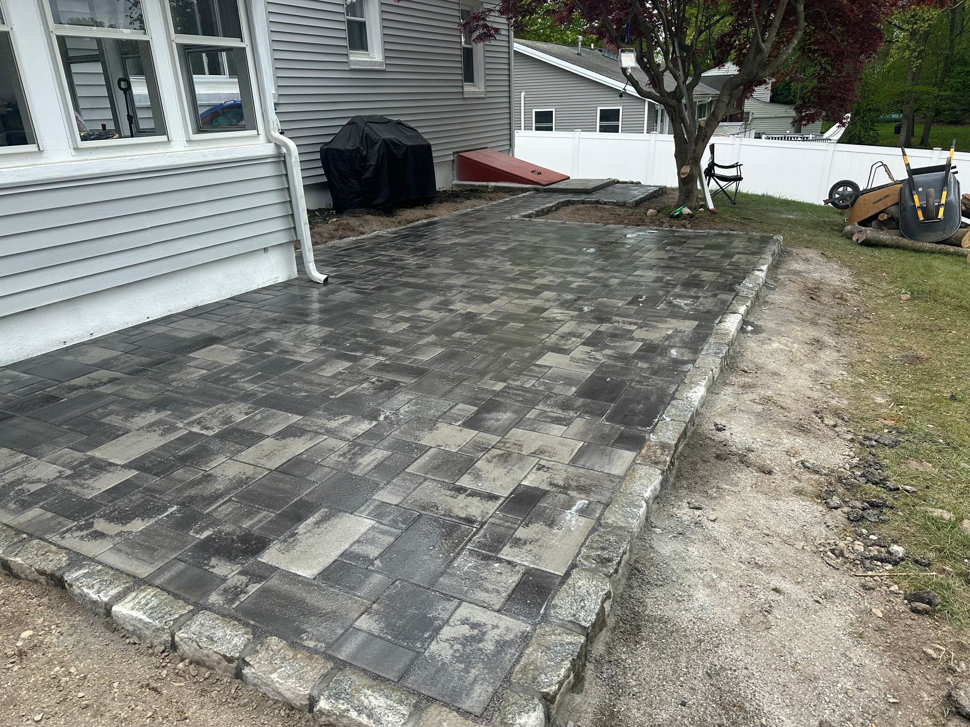 Gray stone patio beside a house, with a grassy edge and yard tools in the background.