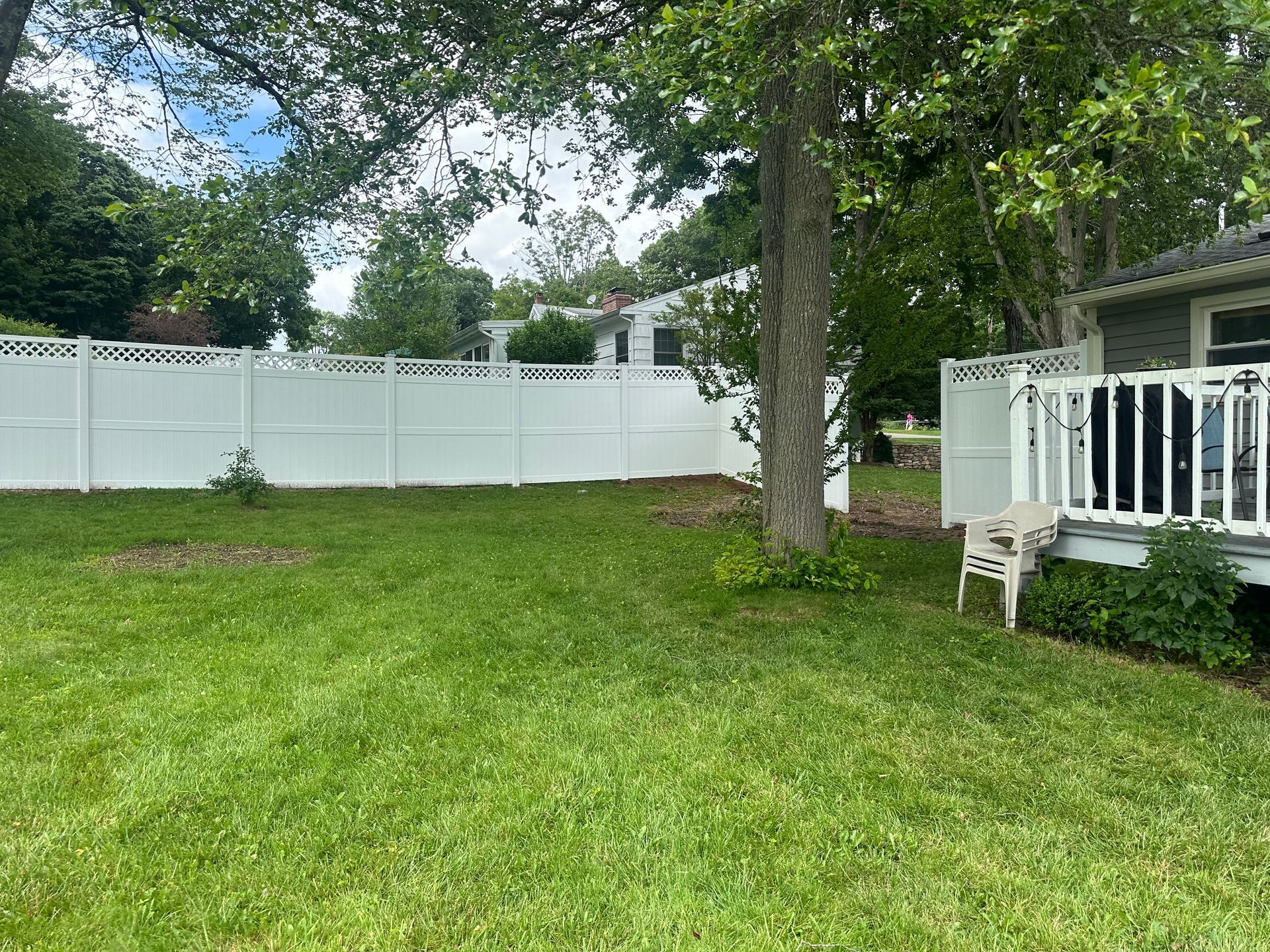Backyard with green grass, white fence, large tree, and a porch with railing.