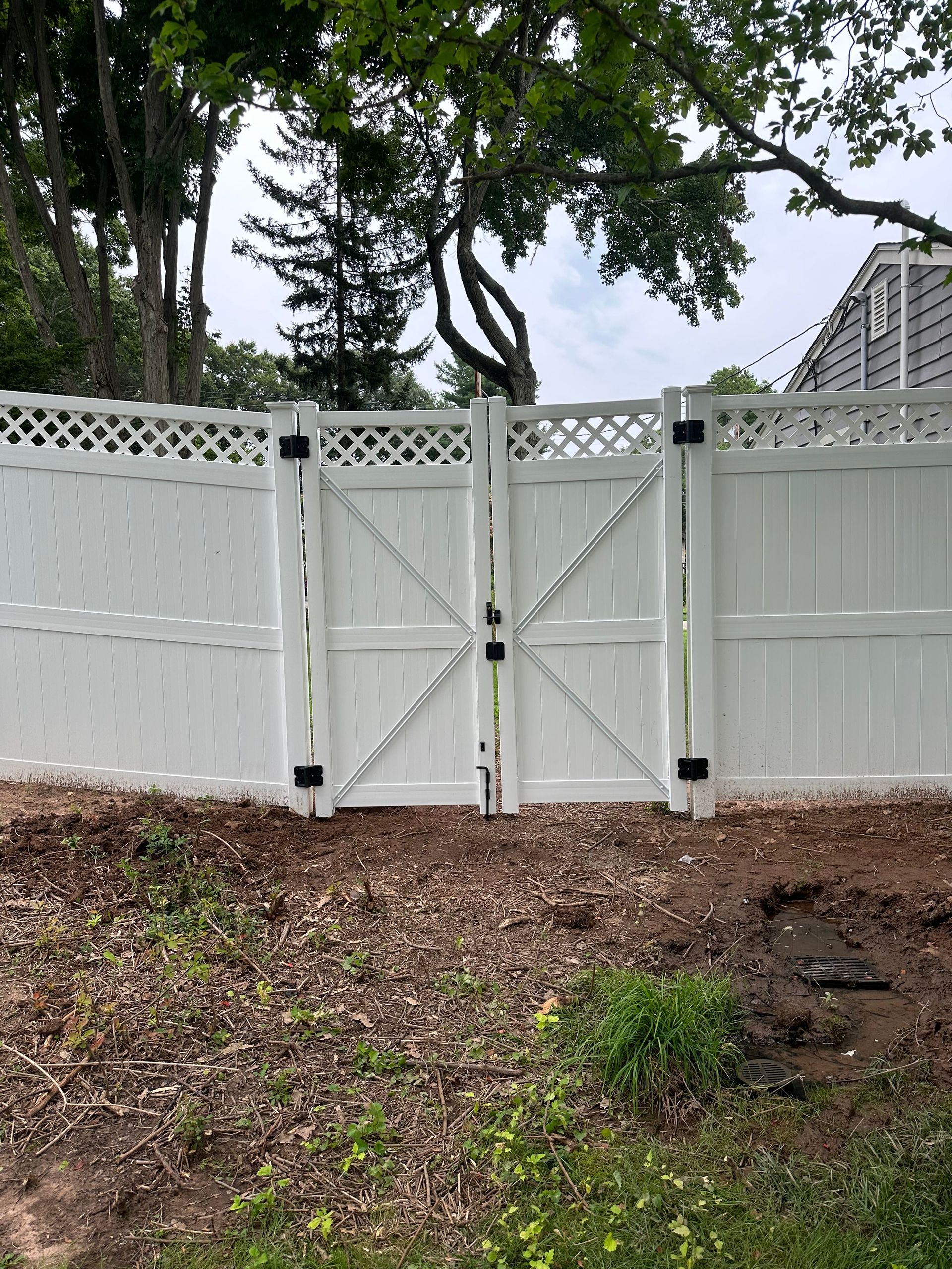 White wooden fence with central gate and lattice panels in a leafy yard, with a hillside and building behind it