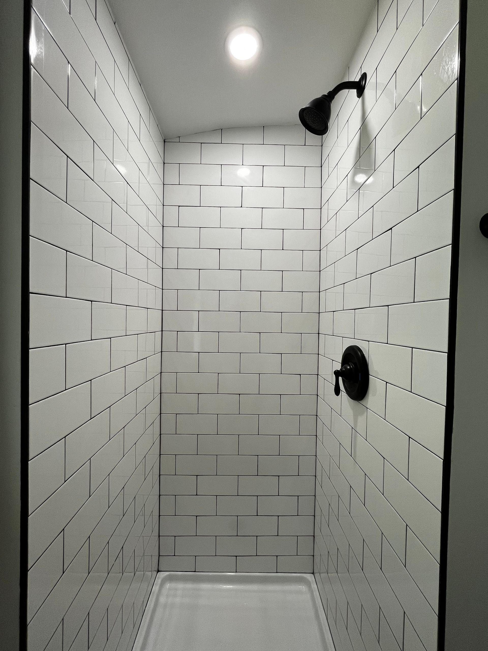 Narrow white-tiled shower with a rainfall head and handheld sprayer under a ceiling light.
