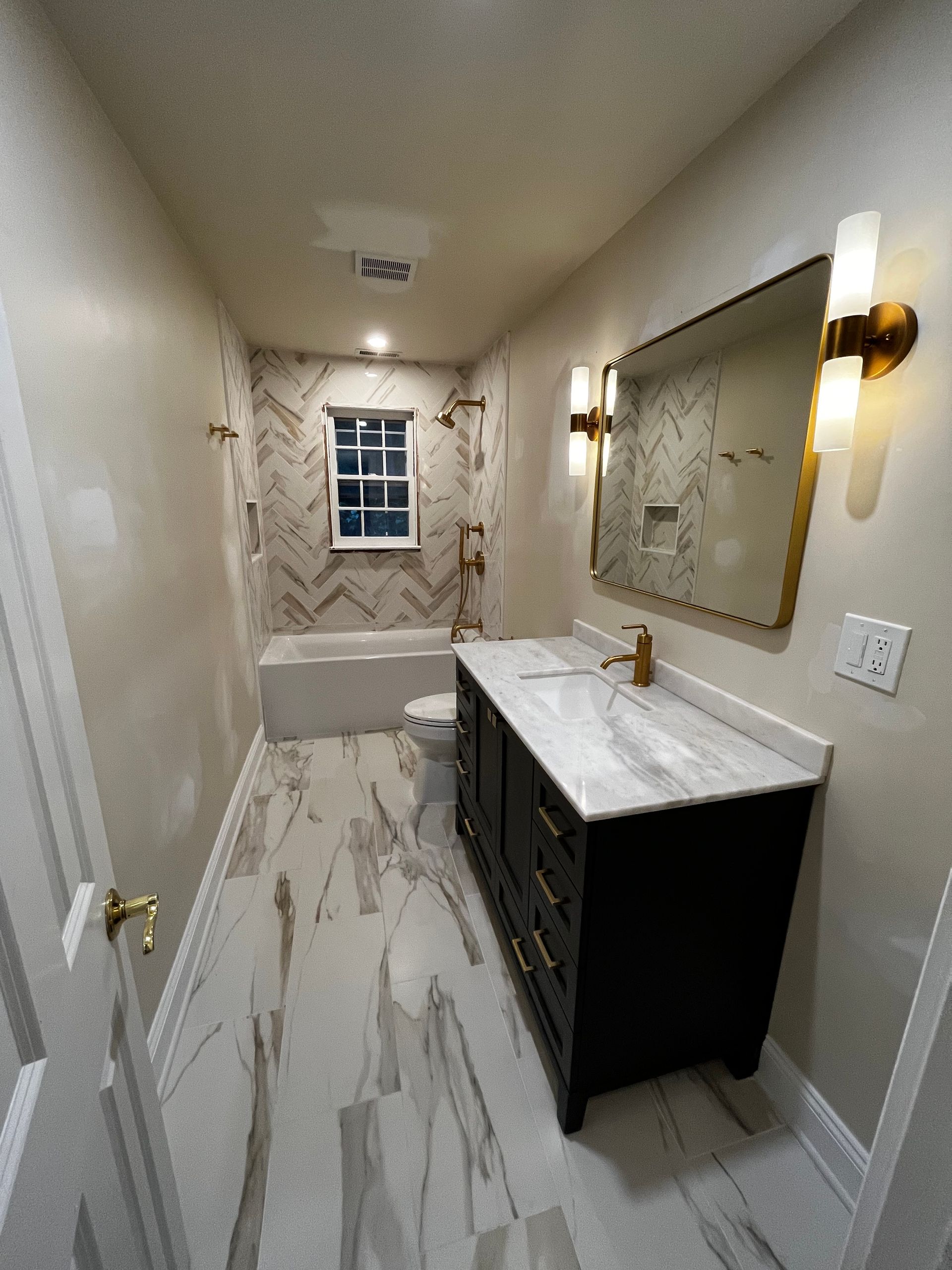 Narrow bathroom with marble tile floor, black vanity, large mirror, tub, and window at the far end