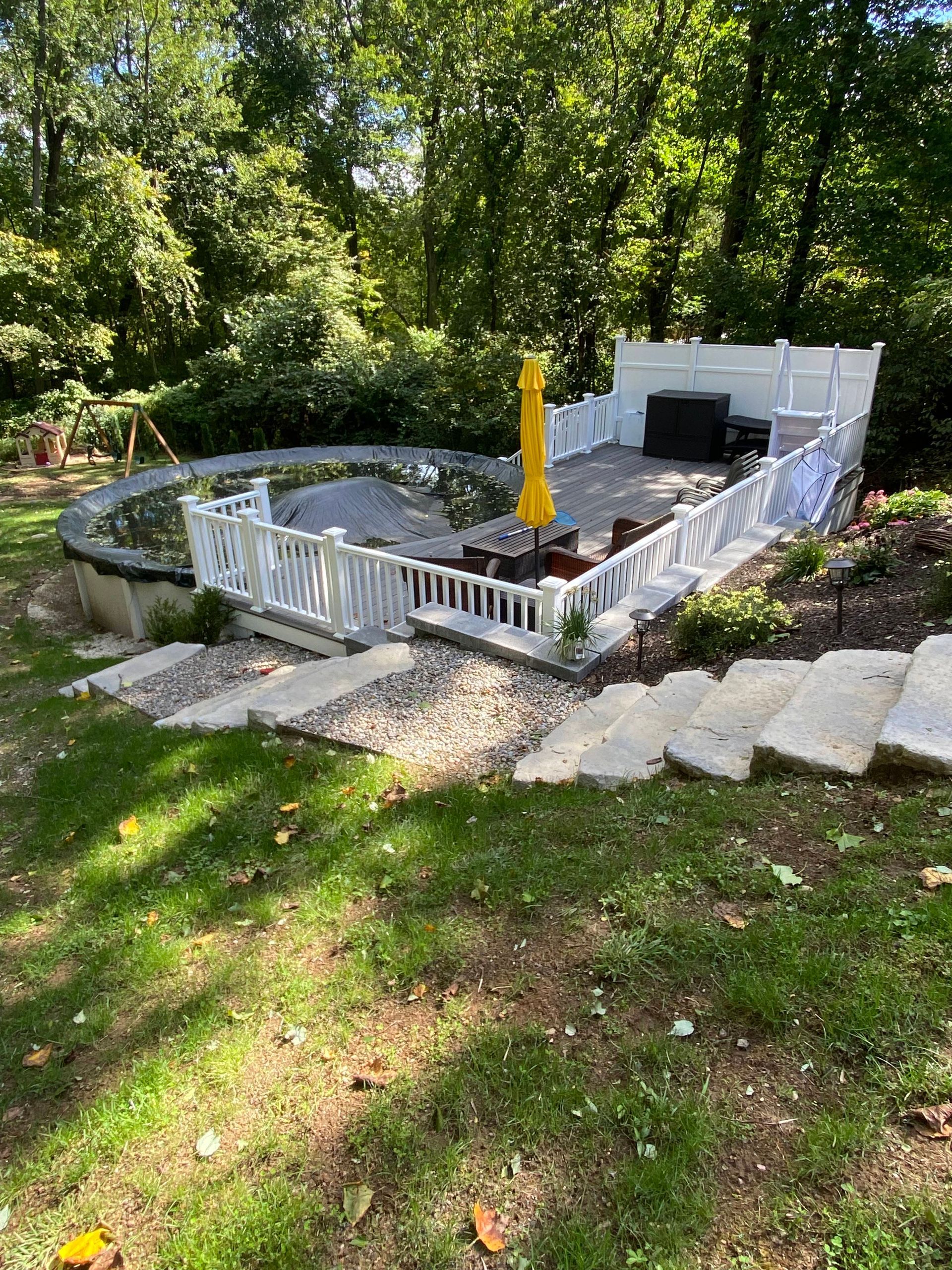Round above-ground pool with white fence and closed yellow umbrella in a wooded backyard.