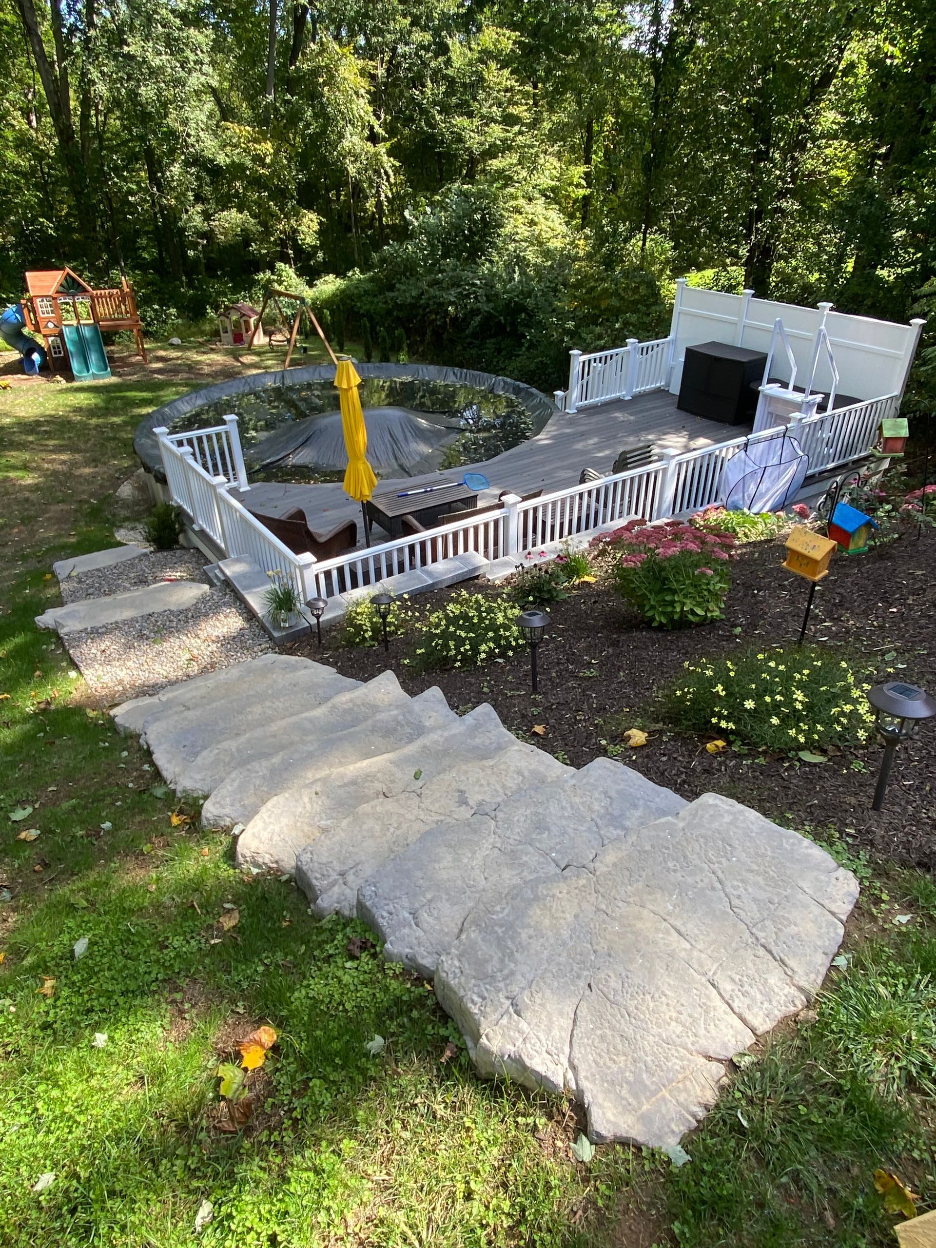 Backyard mini golf-style fenced play area with a yellow umbrella and stone stepping path