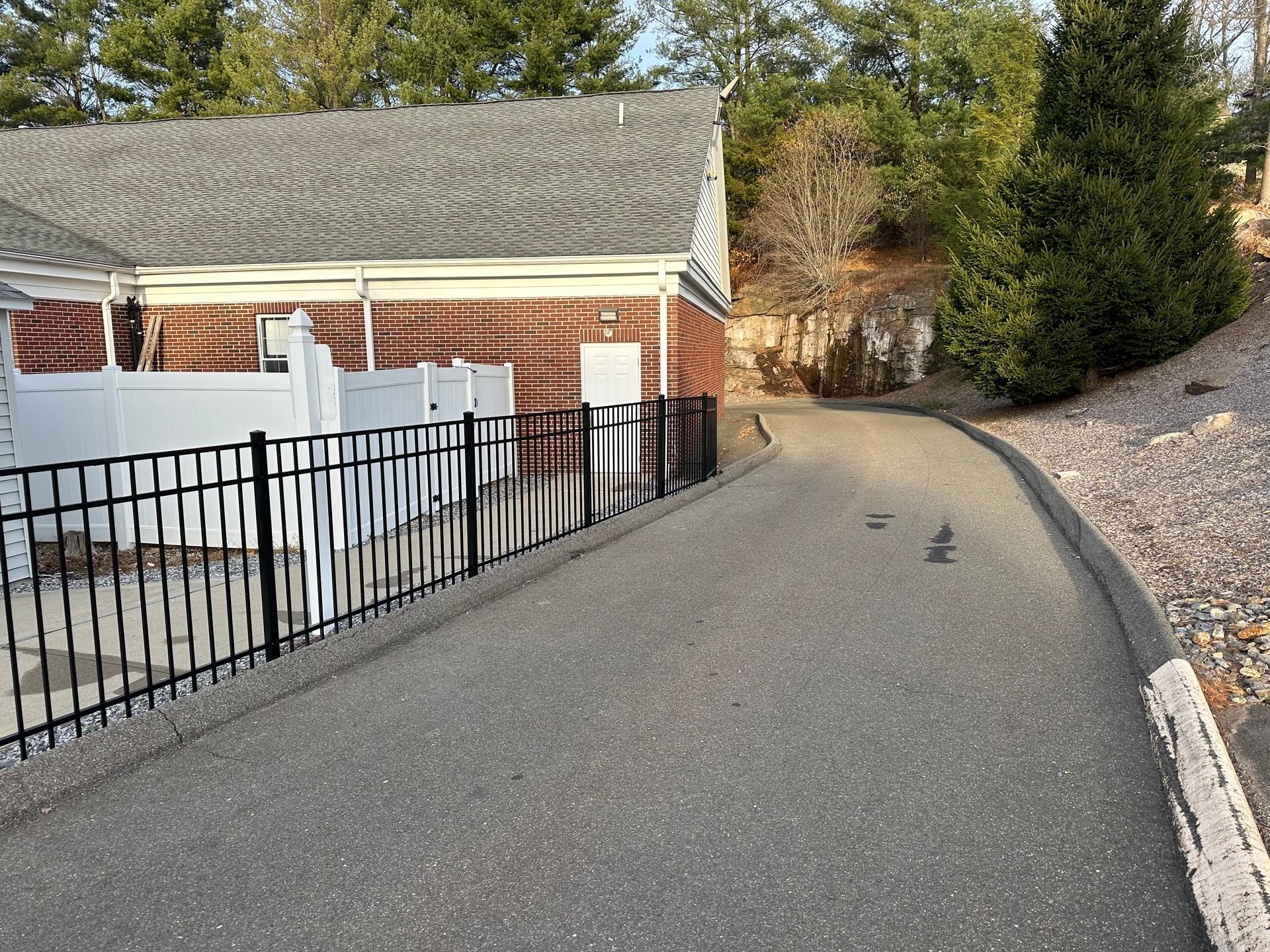 Paved driveway curving beside a brick house, black fence, and gravel embankment with trees