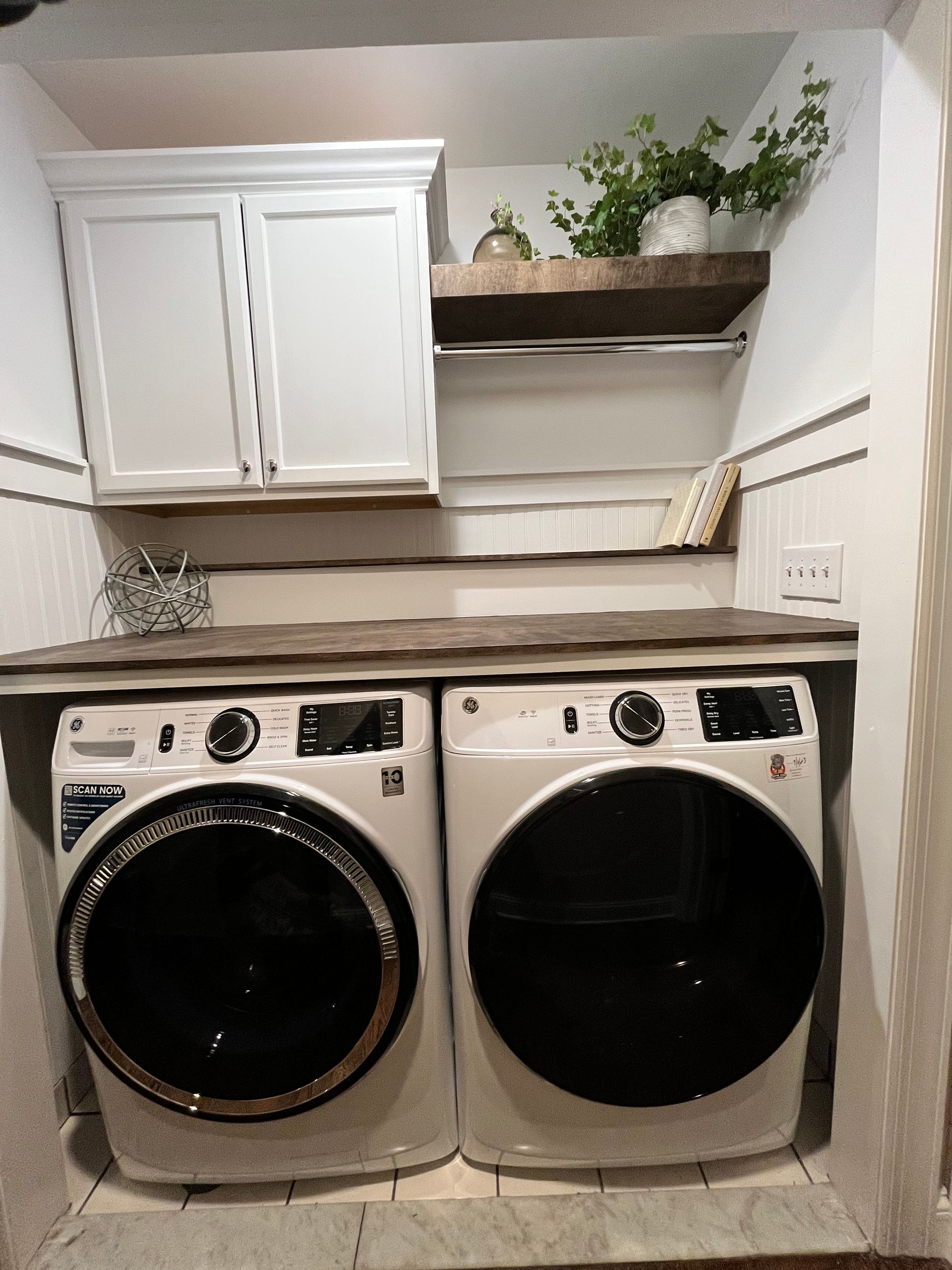 Laundry room with white washer and dryer under cabinets, a shelf, and plants on top.