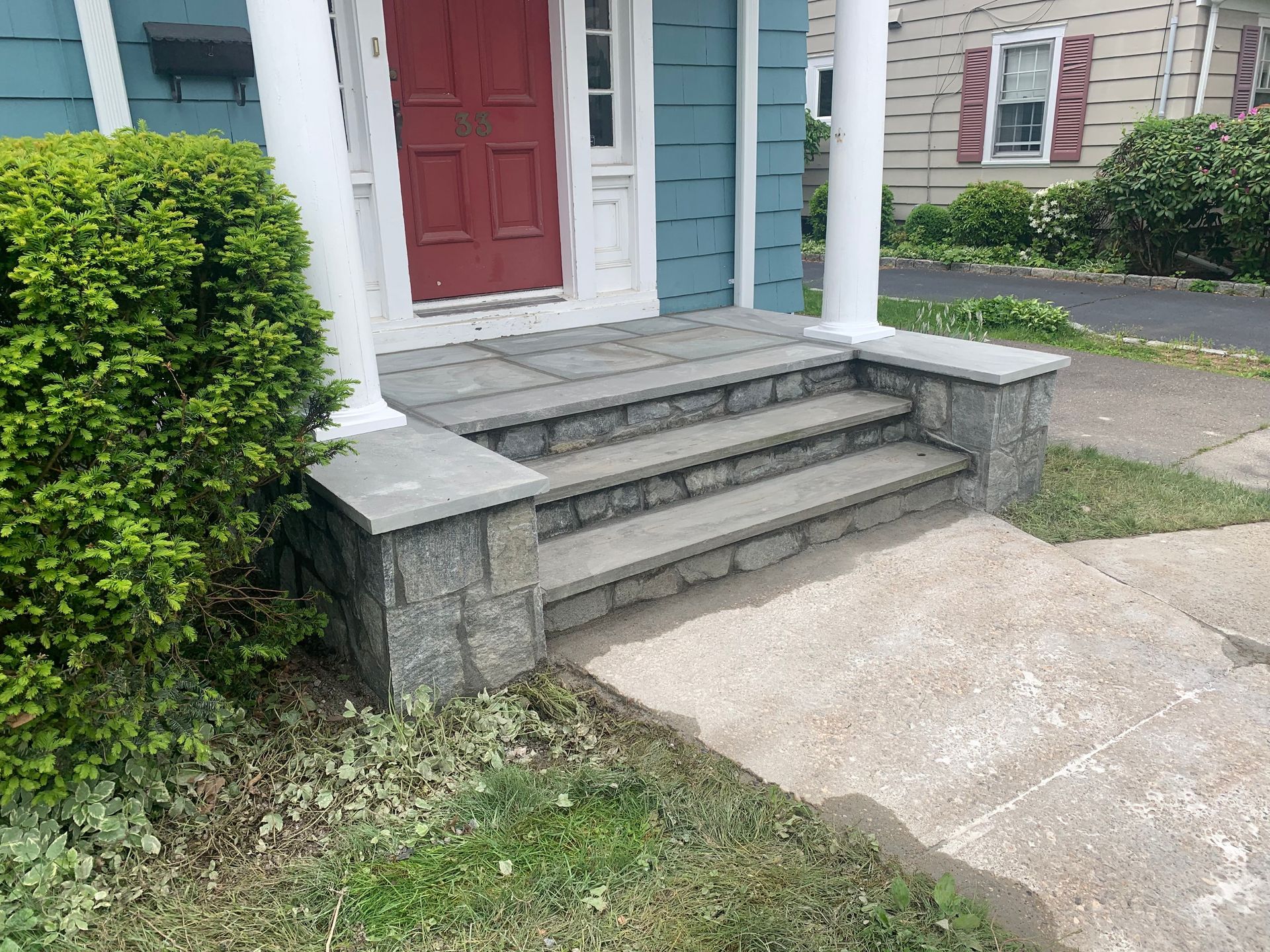 Front porch with red door, stone steps, and concrete walkway beside a hedge.