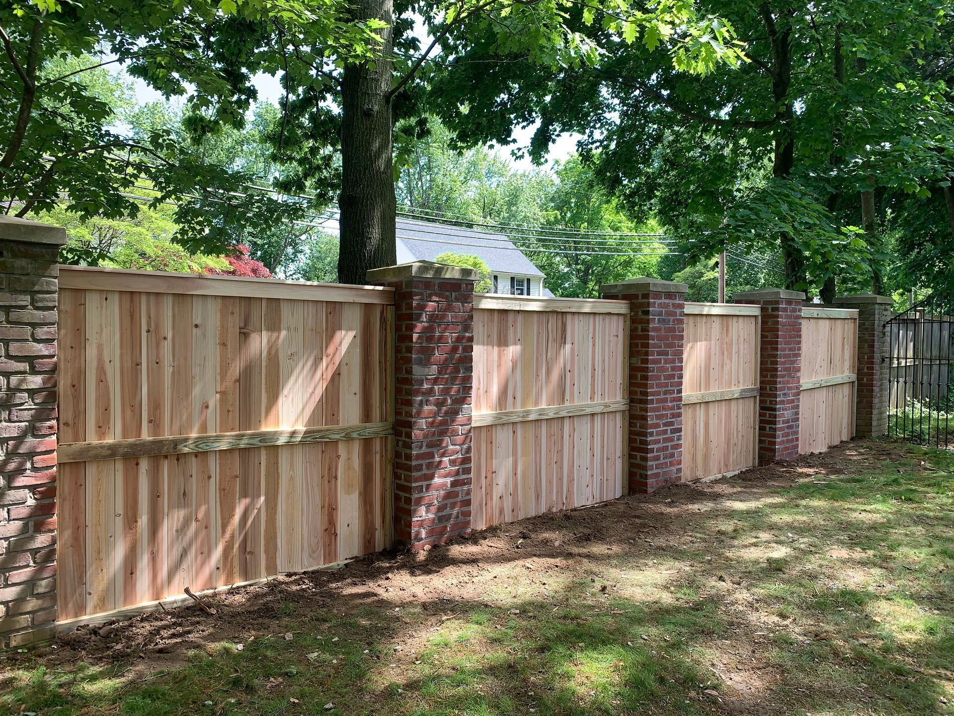 Wooden privacy fence with brick columns along a tree-lined yard, sunlight casting shadows on grass