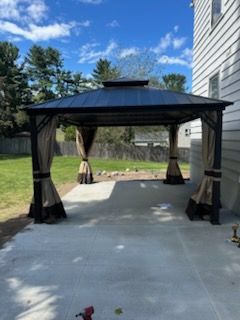Black gazebo with curtains on a concrete patio beside a house and lawn under a blue sky