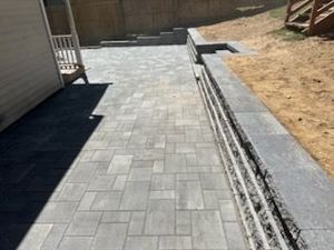 Gray paver walkway beside a house, with a dry dirt strip and retaining wall along the right side.