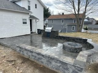 Backyard stone patio with built-in fire pit beside a white house and gray retaining wall