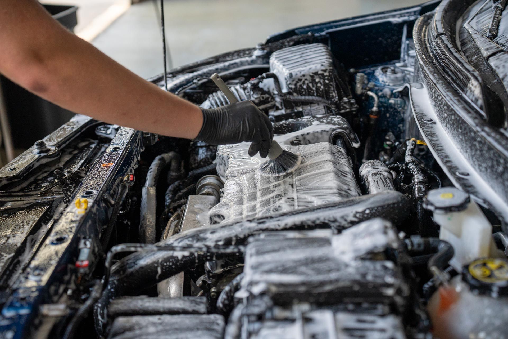 A person wearing a black glove uses a small brush to scrub soapy foam off the engine bay of a car.