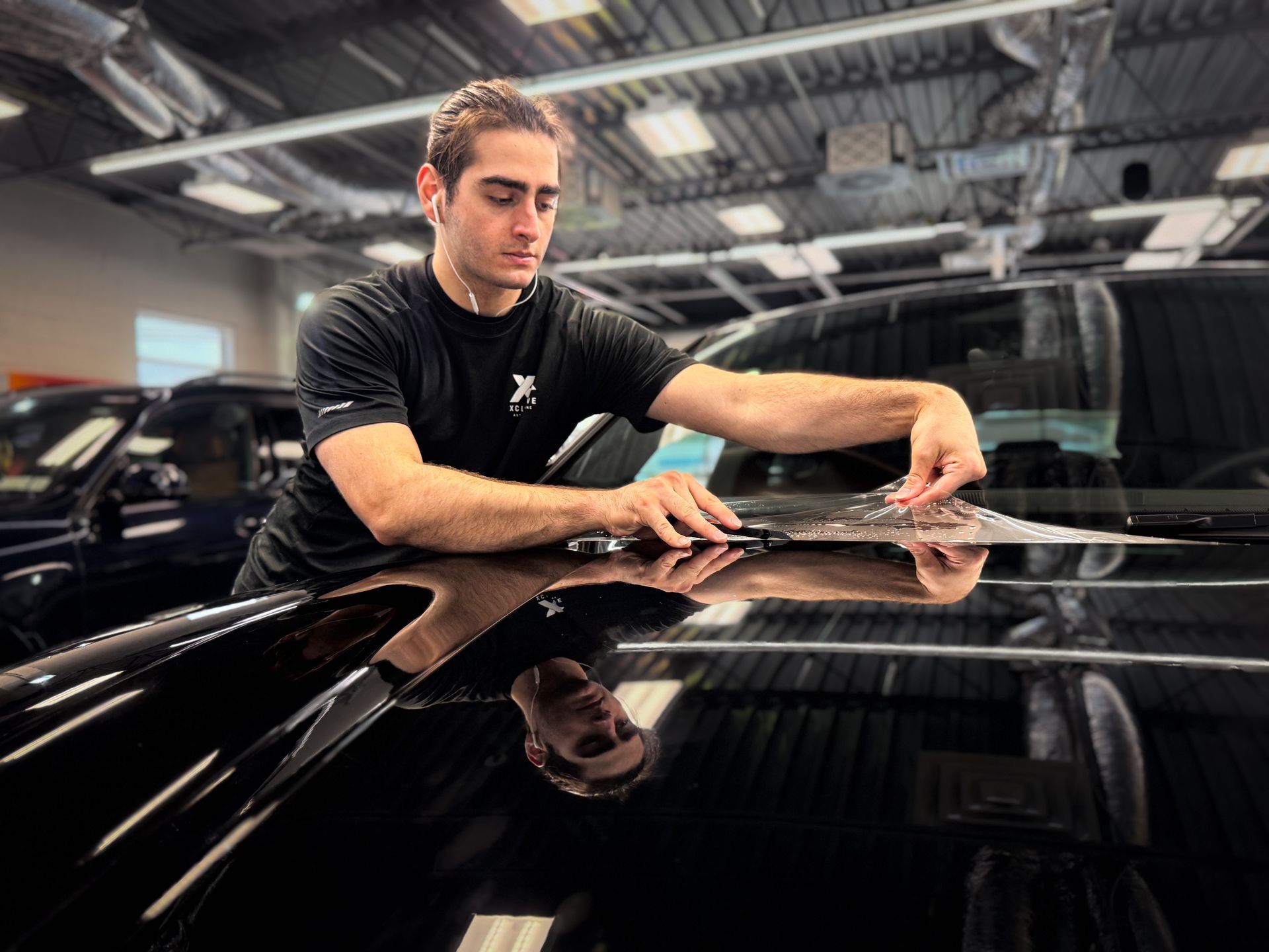 Man polishing a glossy black car in a garage, leaning over the hood under bright lights