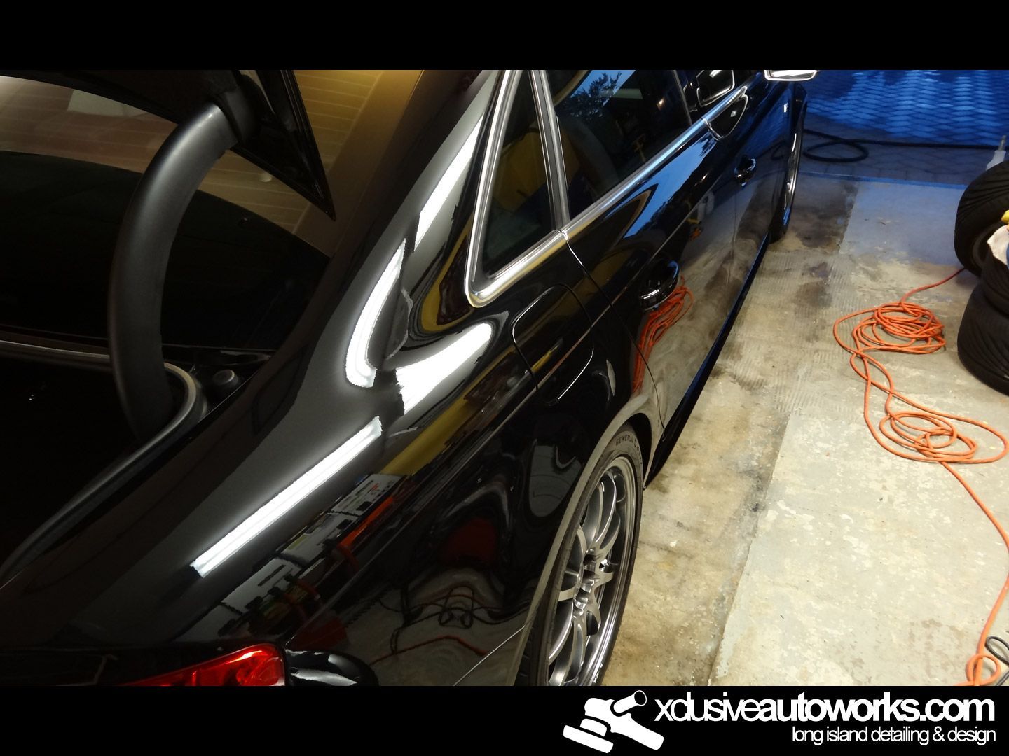 A black car is parked in a garage with the website xdusveautoworks.com in the corner