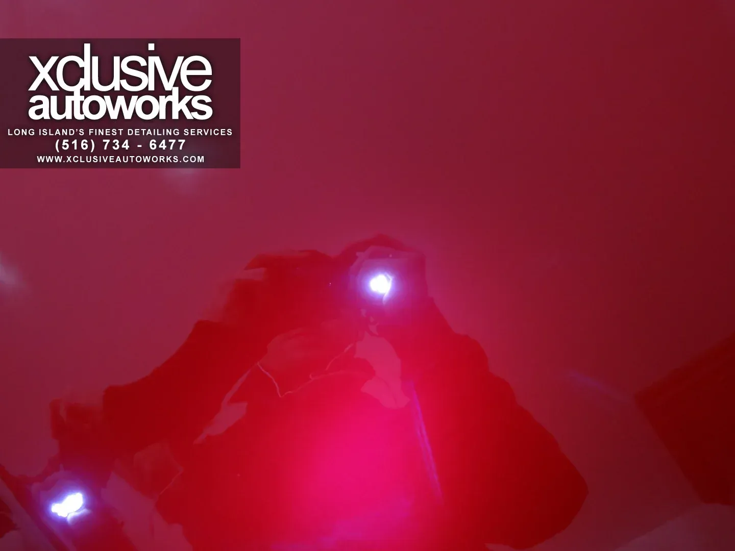 A person taking a picture of a red car with xclusive autoworks written on the bottom