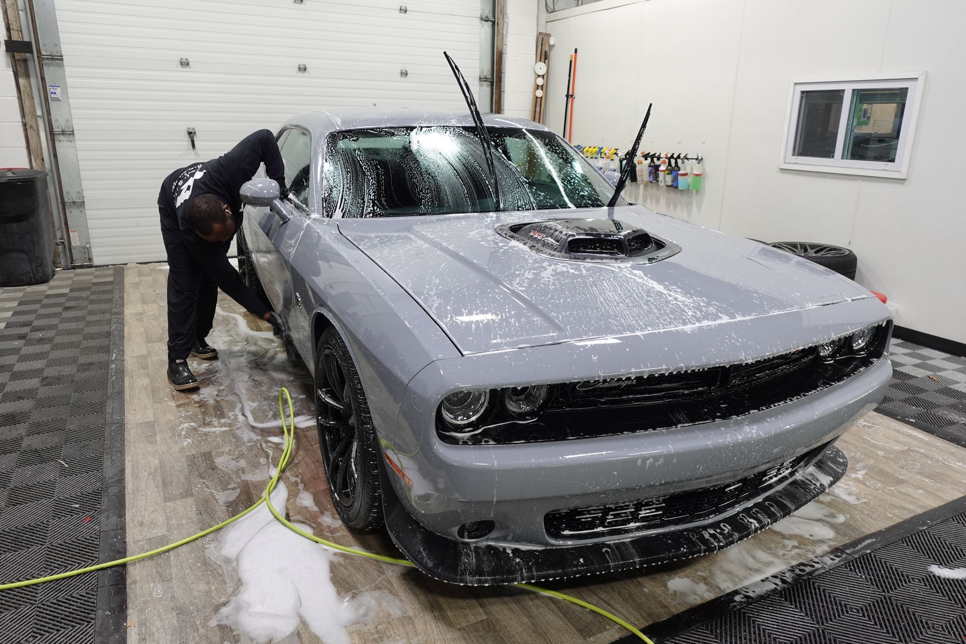 A person washes a gray Dodge Challenger inside a bright, tiled garage.