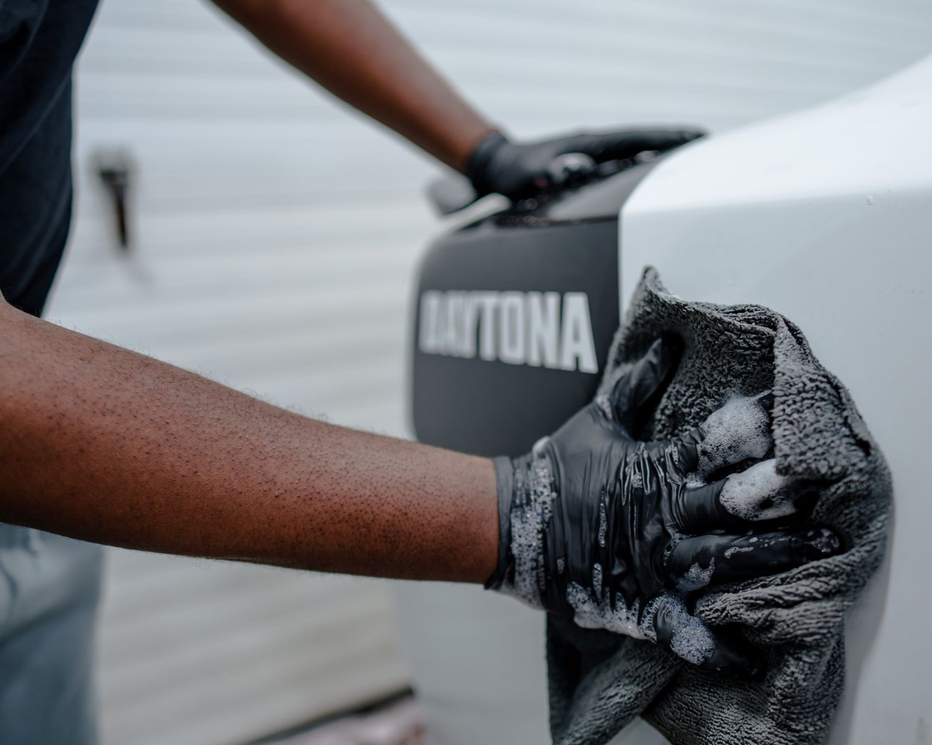 A person wearing black gloves wipes a white vehicle surface featuring a black with a soapy grey cloth.